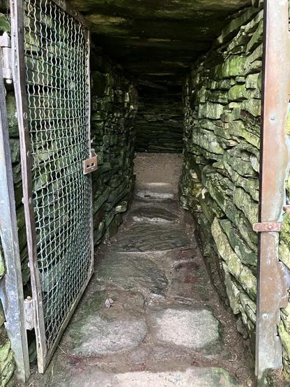 A photo showing the low entrance to the tomb, with mossy stones and a rusted metal gate standing open