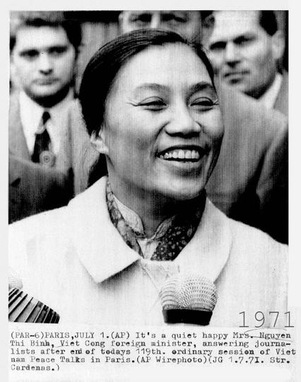Nguyen Thi Binh smiling brightly while holding a microphone, with a blurred background showing several men partially visible. The composition highlights her positive expression and engagement with journalists. Text in the image indicates the date and context of the event as part of the Vietnam Peace Talks in Paris, July 1, 1971. The image has a slightly grainy texture typical of press photographs from that era.
Black and white photo.