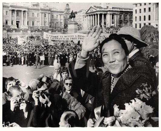 High-contrast black-and-white photograph capturing a rally in an urban square, densely populated with people. The composition centers on Nguyen Thi Binh, prominently positioned in the foreground, waving and smiling while holding a bouquet of flowers. She wears a dark sweater and a patterned scarf. Surrounding her, numerous photographers point their cameras, emphasizing the moment's significance. Behind the crowd, historical buildings are visible, with large banners demanding an end to the Vietnam War, enhancing the rally's political context. The image conveys a sense of activism and public engagement.