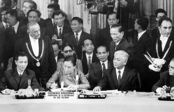 A group of political leaders is gathered in a formal setting, with Nguyen Thi Binh, in a traditional ao dai, signing peace accord at the table. The table displays a nameplate reading "GOUVERNEMENT RÉVOLUTIONNAIRE PROVISOIRE DE LA RÉPUBLIQUE DU SUD-VIETNAM." Surrounding them are various men, some in formal clothing, looking on attentively, while others stand in the background, creating a serious atmosphere.