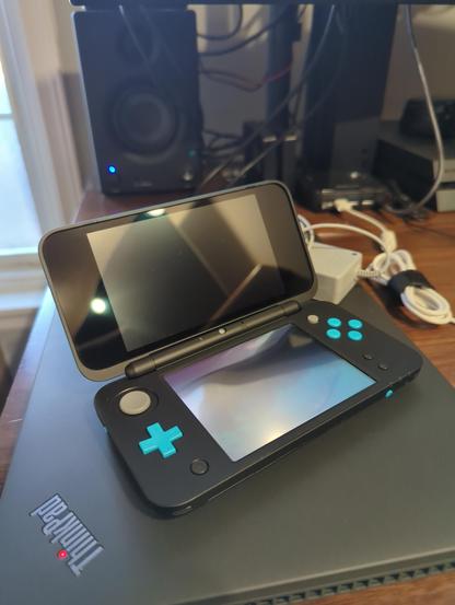 a black and turquoise "new" 2ds XL sitting on top ofathinkpad on a messy desk.