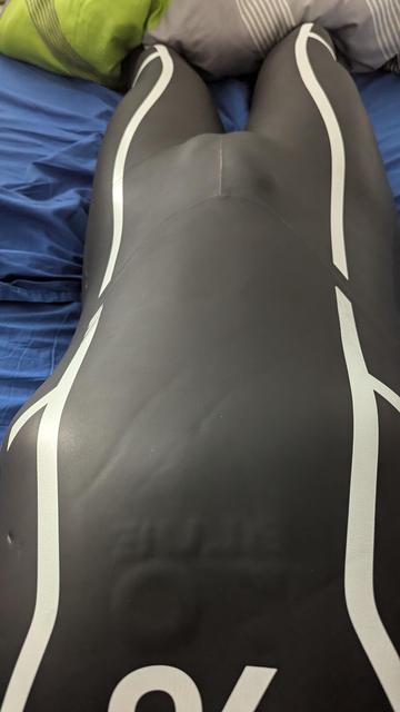 The image shows a person lying on a bed wearing a black neoprene wetsuit with white curved lines running down the legs. The wetsuit fits snugly, highlighting the contours of the body. The material has a smooth, slightly shiny texture, typical of neoprene, and there are a few visible creases in the fabric. The person is lying on their back with their legs together. The bed has blue sheets, and there are pillows in the background, including one that is green with a striped pattern. The perspective of the image is taken from above, looking down at the person’s body.