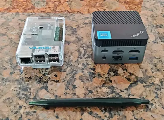 Picture of two mini computer on a kitchen counter top. On the left:  a Raspberry PI 2 on a clear case, it has rectangular shape and it is longer than the other mini pc. On  the right: a GMKTeck, it has a cubic shape and it is double the height of the RPI2.