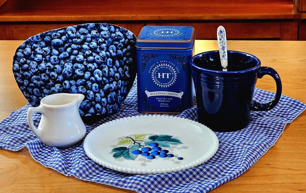A tea tableau in blue and white with blueberry- themed linen & china on a blue gingham cloth, and vintage restaurantware and Fiestaware. The tea is Blueberry Green from Harney and Sons.