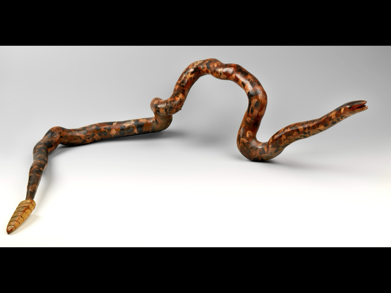 Official museum photo of the object, full profile on offwhite background: twisted wooden root fashioned into a rattlesnake, painted mottled colors of brown, yellow, red, black, carved with an open mouth and embellished with a tongue on one end and a rattle tail on the other; identified as having metal parts but unclear where these may be, might be on either the tongue and/or tail pieces