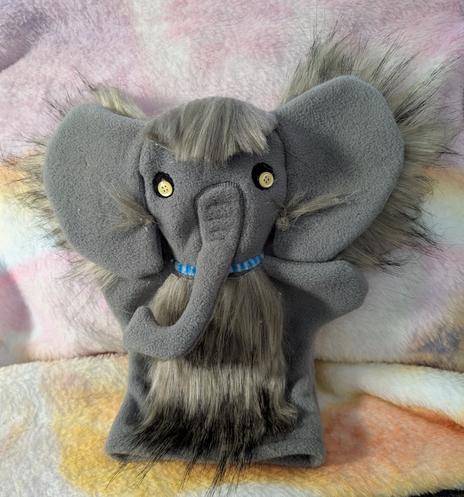 Photo of a grey elephant hand puppet against a pale pink/yellow backdrop. The puppet has been customized with brown faux fur on its head, behind its ears, and on its chest. It also has yellow buttons for eyes, and a blue pipe-cleaner around it's neck.