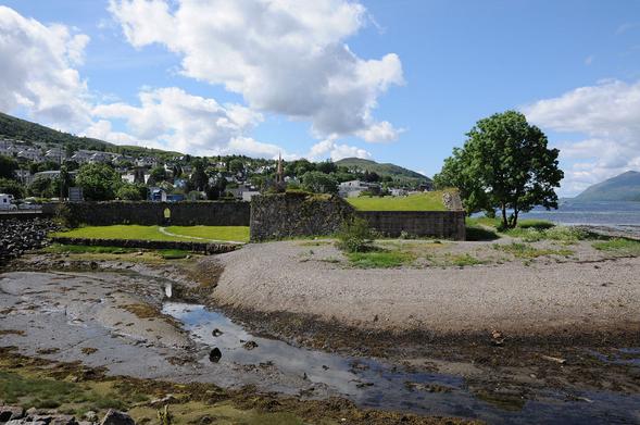 The Old Fort, or Fort William. The image shows a stream in the foreground flowing into the loch at low tide. Beyond it is a shingle embankment and then the walls of the old fort, with a tree standing to their right, between them and the loch visible on the right of the image. The town of Fort William can be seen climbing a hillside in the background.
