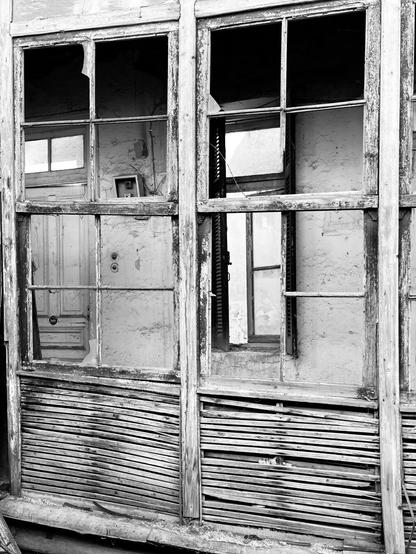 In black and white, the skeleton of a covered porch in a ruined house. The upper part is wooden window frames, almost entirely intact but no glass. Through them, the front door, intact, and a window, with glass and shutters, looking out to emptiness, of the house proper. The lower part of the porch has panels of lathe and plaster, the plaster fallen and the horizontal lines of lathe showing. All the wood is bleached by the sun and weathered, paint stripped away by time. The whole is a composition of horizontal and vertical lines, rectangles and smaller rectangles.