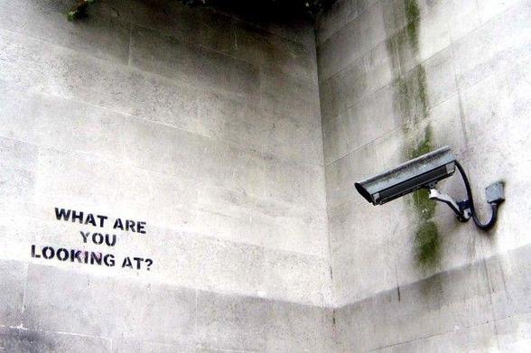 Banksy’s security camera mounted on a stark dirty concrete corner, facing the other wall, which has “What are you looking at?” stenciled on it. Almost monochromatic. Dystopian.