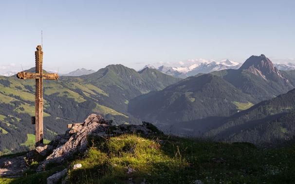 A weathered wooden summit cross stands on the rocky peak of the Gaisberg, overlooking rolling green hills and rugged alpine peaks. In the distance, the prominent Großer Rettenstein and snow-capped mountains add depth to the scenic view near Kirchberg in Tirol, Austria.