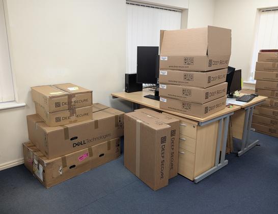 An office desk with no chair, covered and surrounded by large cardboard boxes.