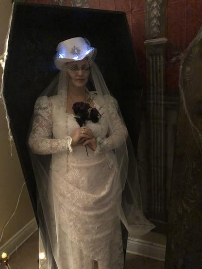 Costume of a ghost bride: woman standing in a coffin with eyes closed, holding dark flowers wearing a white lace over pale pink gown in a late 1800’s style with front draping on the skirt, a sweetheart neckline edged with lace trim and a narrow brimmed hat decorated with lit bats