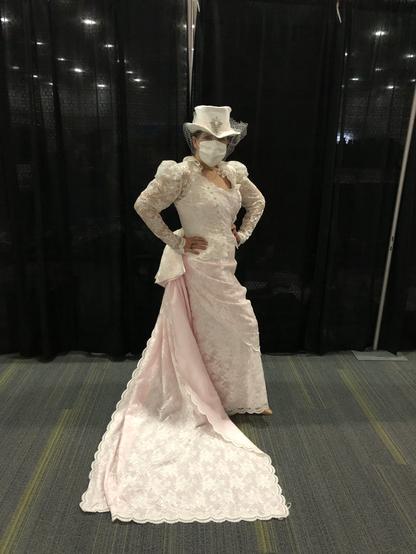 A woman in a bridal gown stands in front of black pipe and drape. Her dress is pale pink polyester shantung with lace overlay and a lace over jacket. The skirt features a draped front apron curving into a bow and 2 metre train in shantung and lace. The jacket has puffed sleeves and a lace ruffle at the back neckline. Her hat and mask are in white satin,