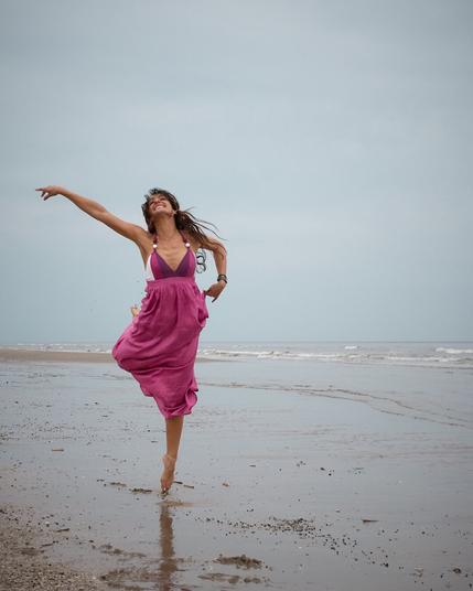 A smiling woman in a purple dress jumping and captured in midair at the beach. In the background the shoreline and the sea are visible during a hazy day.