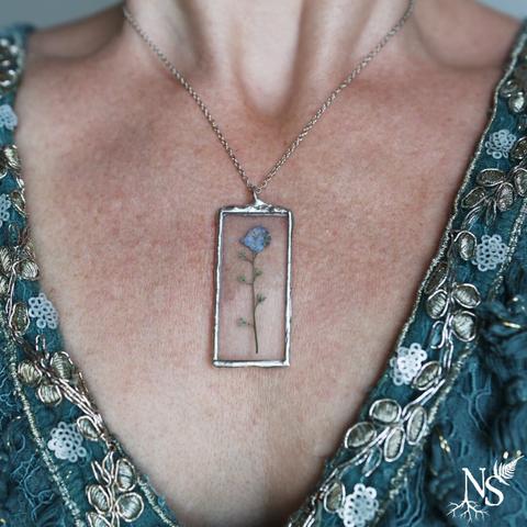 A neckline with a blue dress with silvery shiny lace and a necklace. The necklace is a simple chain and the pendant is made of stained glass and showcases a real pressed forget-me-not flower.
There’s a NS logo in one corner, with roots starting at the bottom of the N and a fern starting at the top of the S.
❉
Un décolleté avec une robe bleue en dentelle brillante argentée et un collier. Le collier est une simple chaîne et le pendentif est en vitrail et met en valeur une vraie fleur de myosotis pressée.
Il y a un logo NS dans un coin, avec des racines commençant en bas du N et une fougère commençant en haut du S.