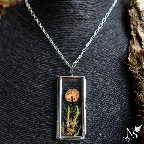 A necklace on a black jewelry display. On the side, you can make out a large piece of bark. The necklace consists of a simple chain and a glass pendant that contains a real mushroom pressed on a bed of pressed moss.
There’s a NS logo in one corner, with roots starting at the bottom of the N and a fern starting at the top of the S.
❉
Un collier sur un présentoir noir. Sur le côté, on devine un grand morceau d’écorce. Le collier est constitué d’une chaîne simple et d’en pendentif en verre qui renferme un véritable champignon pressé sur un lit de mousse pressée.
Il y a un logo NS dans un coin, avec des racines commençant en bas du N et une fougère commençant en haut du S.
