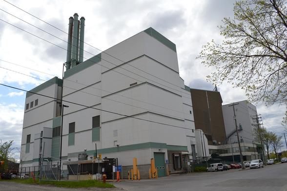 Bâtisse industrielle aux murs gris-beige, d'environ quatre étages. Il y a quatre cheminées peintes en vert qui émergent de con centre. Sur le côté de la bâtisse, il y a une guérite pour faire entrer les véhicules, l'entrée est longée d'une clôture de métal. Il n'y a presque pas de fenêtres sur l'édifice. On peut voir une porte de garage. Plusieurs fils électrique passent devant la bâtisse.
