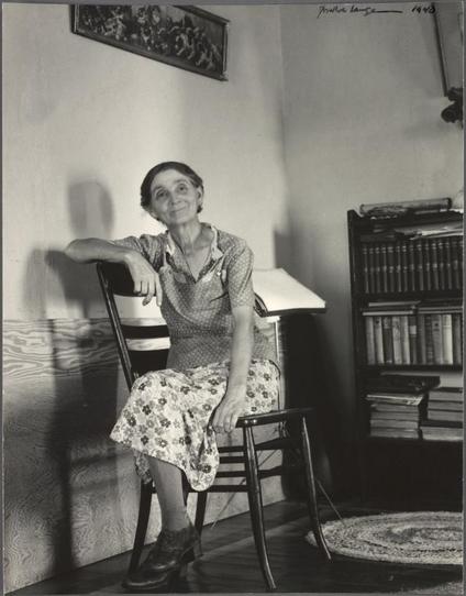 The photograph captures a middle-aged woman seated in an indoor setting, likely her home or farm. She is posed comfortably with one arm resting on the back of a chair and a soft smile on her face. The subject wears a sleeveless blouse adorned with small floral patterns over a matching skirt decorated with large flowers. Her shoes are simple yet sturdy, suitable for working outdoors.
Behind her sits an open bookcase filled to capacity with numerous books, indicating a well-read individual or someone who values knowledge and education. To the left of the woman stands a traditional desk lamp, its light illuminating part of the scene. The background wall is adorned with what appears to be framed artwork depicting rural scenes, contributing to an atmosphere reflecting her life on the Great Plains in Nebraska.
The monochromatic tones of this black-and-white photo suggest it was taken during or shortly after World War II (1943), as indicated by a handwritten inscription at the top. The image exudes warmth and tells a story about the subject's daily life, work, and surroundings on her farm amidst challenging times in America's history.