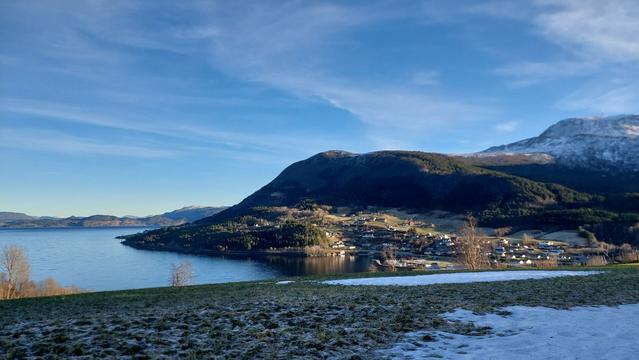 In the foreground a green, frosty field with patches of snow. 
To the left the fjord, and then a bay and a village with houses up the hill towards a round, tree covered mountain. In the background to the right a higher mountain that looks dark blue and is partly covered in snow. The sky is blue with a few wispy clouds. Flat sunlight is coming in from the right on the village and mountains on the other side of the bay.