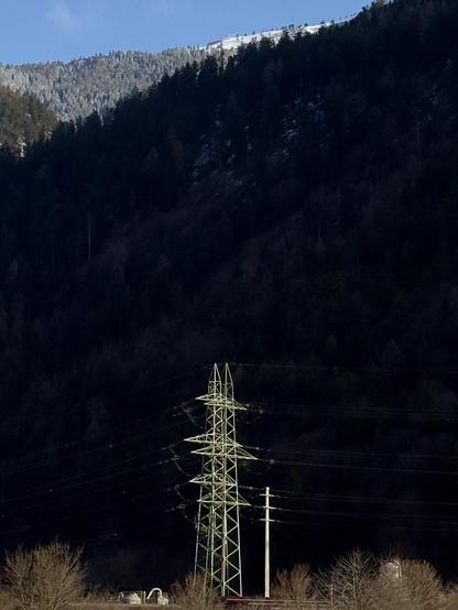Zwei von der Sonne angestrahlte Starkstrommasten stehen vor einem schattigen Berghang. Oben drüber sieht man noch knapp den blauen Himmel, einen verschneiten Wald und eine verschneite Alp.
