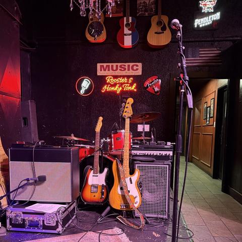 A Fender Telecaster and P-Bass waiting on stage in guitar stands next to their respective amplifiers in front of a red drum set. Three guitars hang on the wall behind them with a Roosters Honky Tonk neon sign below.