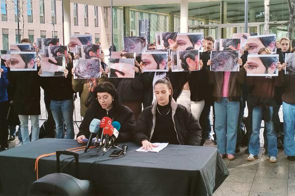 Roda de premsa. Dues persones parlant amb micros en una taula instal·lara davant la Ciutat de la Justícia. Darrera, to de gent sostenint fotos de persones amb lesions