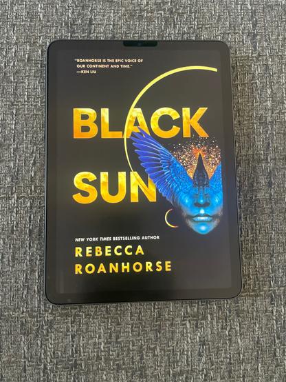 On a black background, a blue human face merges from the nose upwards into a pair of crow’s wings and a crow’s head where the person’s forehead would be. A constellation of stars is around the crow’s head. An eclipsed sun shines around the person’s head, with the title BLACK SUN written in yellow on the edge of the eclipsed sun.