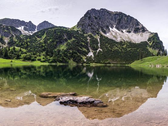 A crystal-clear alpine lake, the Lower Gaisalpsee, reflects the surrounding rocky peaks and lush greenery. The lake is framed by a serene mountainous landscape near Oberstdorf in the Allgäu region, Germany.