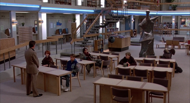 La imagen muestra el interior de una biblioteca moderna y espaciosa. Hay varios estudiantes sentados en mesas de estudio, algunos trabajando en sus libros y otros conversando. En el fondo, se ven estanterías llenas de libros, una escalera y un catálogo de tarjetas.  Una escultura abstracta de bronce se destaca en el centro de la sala. El ambiente es tranquilo y ordenado.  Un profesor o bibliotecario habla con una estudiante cerca de un mostrador de información.