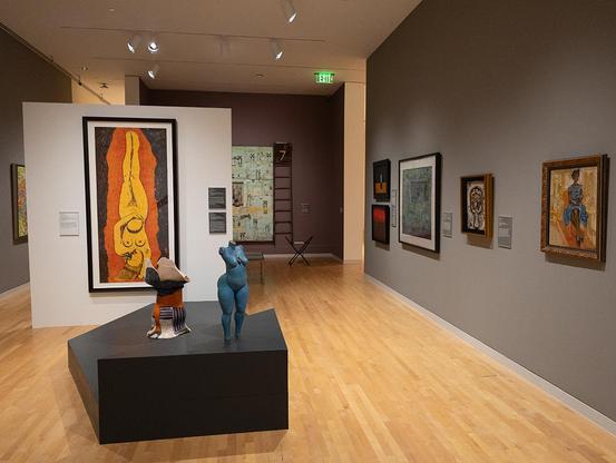 A color photograph of the newly installed Boone and George-Ann Knox Gallery II within the permanent collection of the Georgia Museum of Art at the University of Georgia. Two sculptures (a nude body in blue by Donald Locke and a ceramic chair by Camille Billops) stand on a geometric black pedestal. A large print by Alison Saar of an upside-down nude woman hangs on the wall behind them.