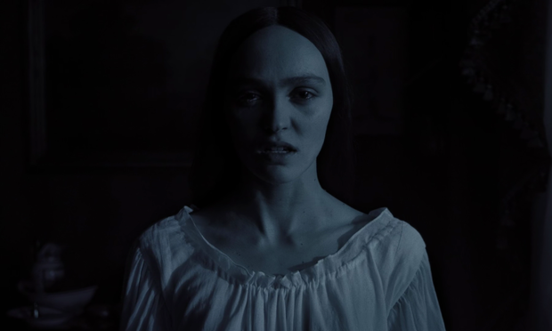 Una mujer joven de cabello oscuro, con una expresión seria y ligeramente angustiada, está en primer plano. Lleva un vestido blanco sencillo de estilo antiguo. El fondo está oscuro y apenas iluminado, creando una atmósfera sombría y misteriosa. La imagen está en tonos azules y grises, acentuando el ambiente melancólico.