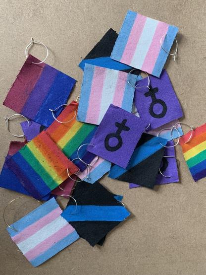 O fondo é dunha sorte de contrachapado. Enriba hai espaiadas varias “bandeiras” de causas sociais (en formato brinco): feminista, lgtbiq+, trans, bi e movemento nunca máis. Vense como anacos de tea pintados.
Fin da descrición.