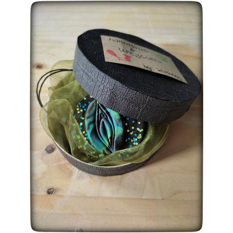 Man sieht eine runde Geschenkbox, mit schwarzem Crocoprint beklebt. Der Deckel ist halb geöffnet. Im Inneren ist ein Vulva-Amulet auf grünem Stoff zu sehen.