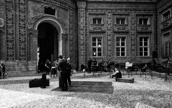 The image depicts an outdoor scene in what appears to be an ornate courtyard with intricate architectural details on the brick building. There are people gathered, possibly setting up or rehearsing, with music stands and equipment visible. The setting suggests preparations for a performance or event. It's a black and white photograph.