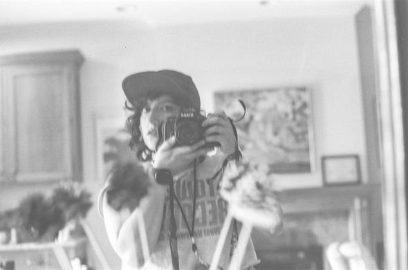 For those who can't see it's a black and white 35mm mirror selfie. You can see in the mirror I'm using a nikon n90. Wearing a Tower Records Tshirt and a wide brimmed baseball/trucker style cap. You can't see this but it's a Glacier national park cap, see you get some information the sighted audience didn't,  and also I have a credential around my neck, which the sighted audience doesnt know is actually from pitchfork music festival 2021. In the background is a mantel with some art above it. This was my childhood home which my father lost last year. Trying to give little extra moments in these alt texts. Hope you appreciate it.