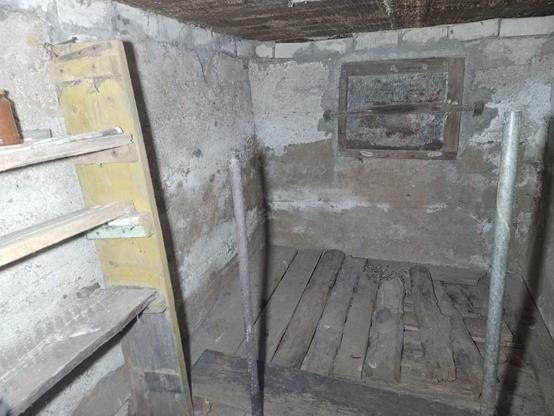 Part of a basement. There's some wooden boards where the potatoes used to be stored. Two steel pipes cemented into the floor to hold retaining boards. A wooden shelf on the left holds a glass jar with yellow stuff inside. The far wall has a steel mesh in a wooden frame covering the potato chute.
