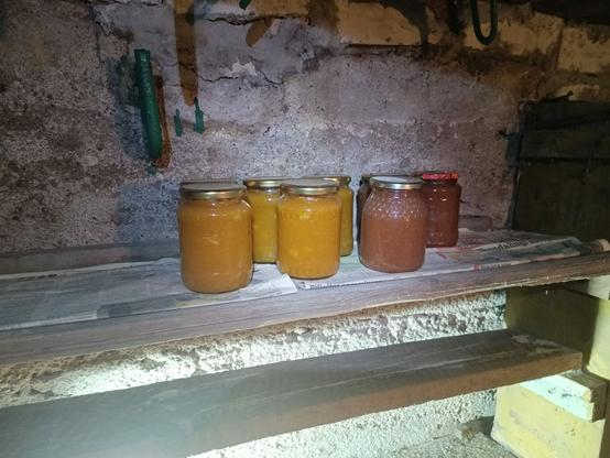 A couple jars of mystery preserves, some yellow, some reddish, sitting on a shelf.