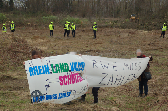 Aktivist*innen stehen an der Mahnwache mit Protest-Bannern: "Rhein.Land.Wasser.schützen." und "RWE muss zahlen". Im Hintergrund steht eine Reihe RWE-Werkschutz mit Bauhelmen und gelben Warnwesten am Waldrand.
