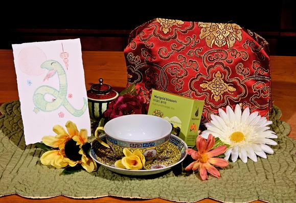 A tea tableau to honor Tết Nguyên Đán Year of the Snake, with lucky red and gold and flowers, and a drawing of a friendly snake under a lantern. The tea is Marigold Blossom herbal.