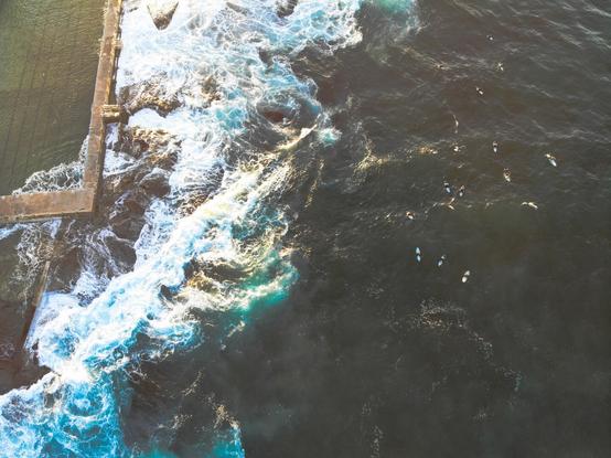 Waves crash against a rock-pool in Sydney's Northern Beaches while surfers sit adjacent. Shot from above with a drone.
