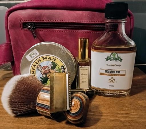 (ChatGPT description) 

The image showcases a classic wet shaving and grooming set. It includes a multicolored wooden-handled shaving brush, a brass safety razor, and grooming products like Stirling Soap Co. Mountain Man Aftershave Splash, Mountain Man shaving soap, and Atelier Rebul Istanbul Eau de Parfum. A burgundy leather toiletry bag sits in the background, adding to the vintage, refined aesthetic.