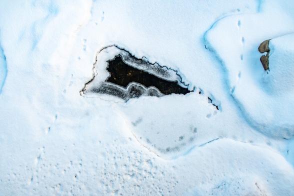 A snow covered mostly frozen river with a triangle shape of near the center of that is clear of snow and ice. In the snow there are tracks