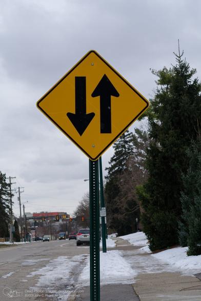 A photograph of yellow diamond-shaped traffic sign which indicates two-way traffic flow. The sign is mounted on a metal post along a city street, with cars visible in the background. The image portrays an urban street scene in winter, with snow on the ground and sidewalks. Against the dark areas of the frame, falling snow is just visible.
