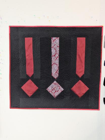 A small wall quilt, slightly wider than it is tall. There are 3 red blocks that look like exclamation points, on a black background. They are arranged in a row, left to right. Instead of a "point" or dot at the bottom, there is a square arranged like a diamond, with the corners facing up/down and left/right. The red pieces are made from thrifted silk ties, and the black background is cotton fabric from my stash. It is bound in red cotton fabric. The quilting pattern is all-over "swirl chain", which features interlocking spirals.