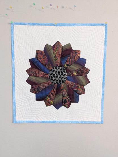 A square-ish mini-quilt with a single Dresden Plate block centered. The Plate block measures about 15 inches (38 cm) across, and is made from 3 silk neckties. There are 18 blades in the Plate, arranged in a circle such that they are radiating outward, ending in points at the outermost edge. They alternate between a fancy redish print and an elegant blue, red, and gold stripe pattern. The center of the Plate holds a circle (about 4.5 inches (11 cm) across, which is made from a black and silver diamond print silk tie. The white background and blue binding are cotton fabrics from my stash. The overall size is about 21 inches (53 cm) square, more or less.