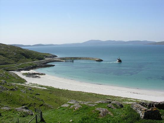 An idyllic hebridean view over the Sound of Barra. The image shows a view down onto a curving white beach in the foreground with blue and turquoise seas. There's a small harbour on a rocky headland on the left of the picture and a ferry is just departing. On the horizon are a series of islands with low hills. The sky is clear blue.