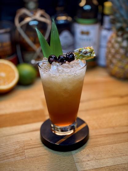 brown colored cocktail with pineapple fronds, three maraschino cherries and a pineapple chunk garnish on a metal cocktail pick sitting on a black and gold coaster with bottles of rum, falernum, pimento dram, and angostura bitters with citrus and a pineapple in the background