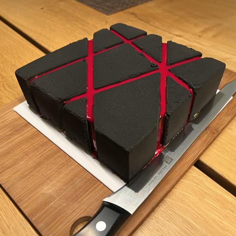geometric square mousse cake with diagonal lines engraved into its surface, sprayed with molten black-colored cocoa butter and red mirror glaze in the grooves. The cake is placed on a white cake board, on a wooden cutting board, and a long knife with a thin blade rests next to it.