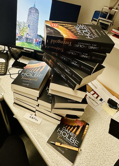 Bücherstapel des Krimis "Totengold". Im Hintegrund das Bild eines historischen Turmes.