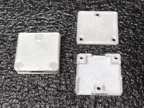 Three white 3d printed parts on a black background. On the top right is the flat rectangular base with 3 mounting holes, under this is the top of the rectangular case which has breaks in the perimeter for the USB connectors and 3 mounting holes. To the left is the assembled case with a PCB sandwiched in between the two parts held together with 3 white nylon screw/nuts.